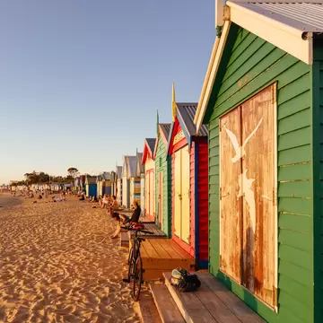 People sitting along the colorful Brighton Beach bathing boxes at Denby St beach and golden sand. 