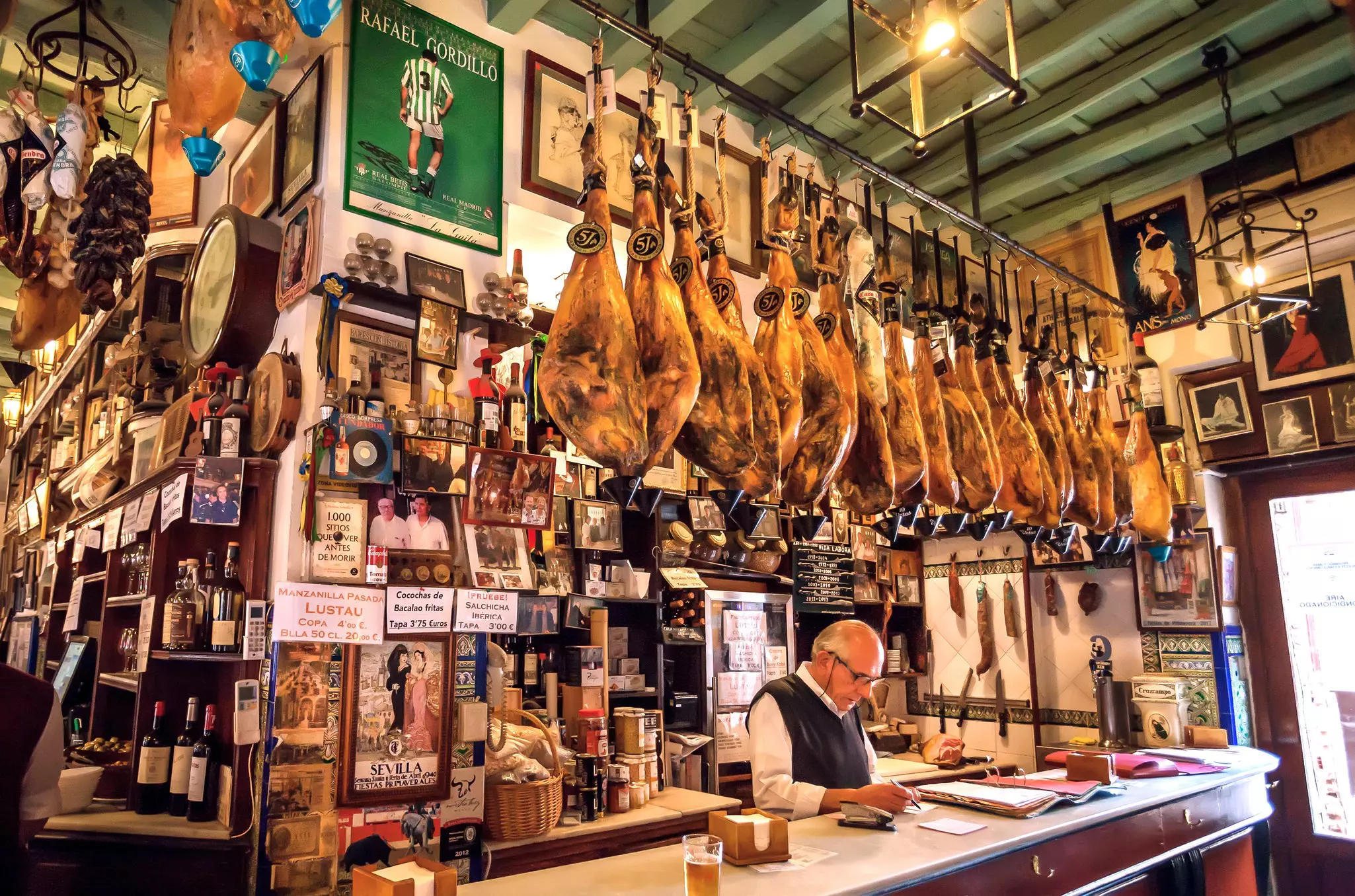 A man stands behind a bar. Dried hams hang overhead, and the wall behind him is filled with photos, signs and bottles.