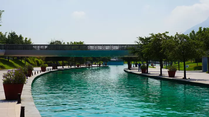 A green river sparkles in the sun lined by a cement walkway and landscaped trees a covered bridge crosses it.