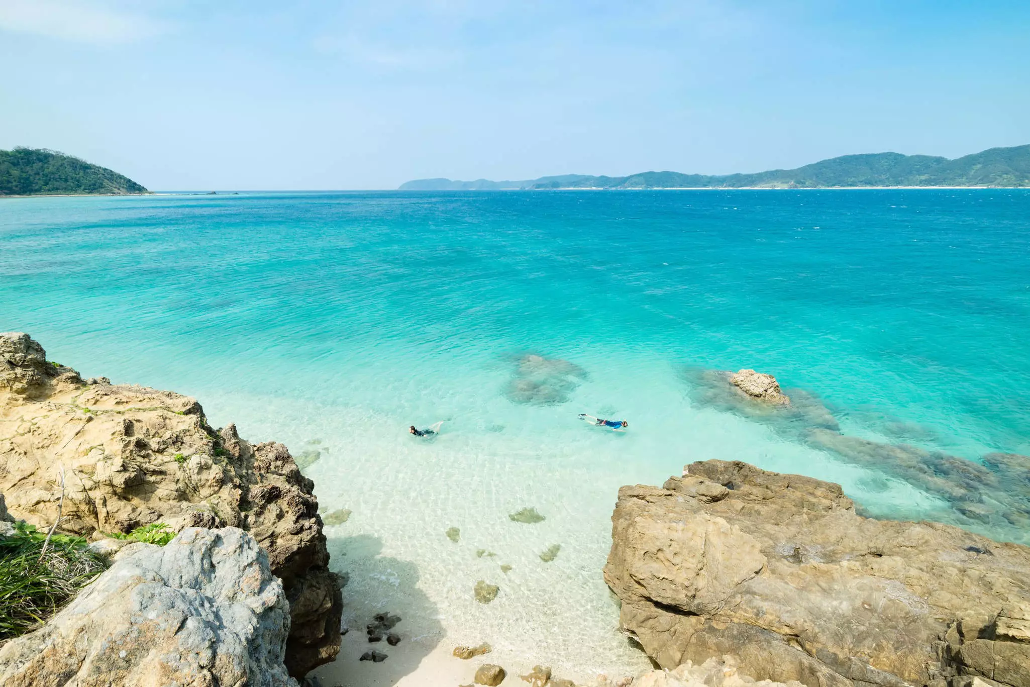 Two snorkelers explore crystal-clear ocean near a rocky shoreline.