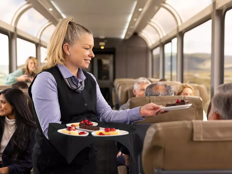 A woman with a blonde ponytail serves desserts on a plate to train passengers.