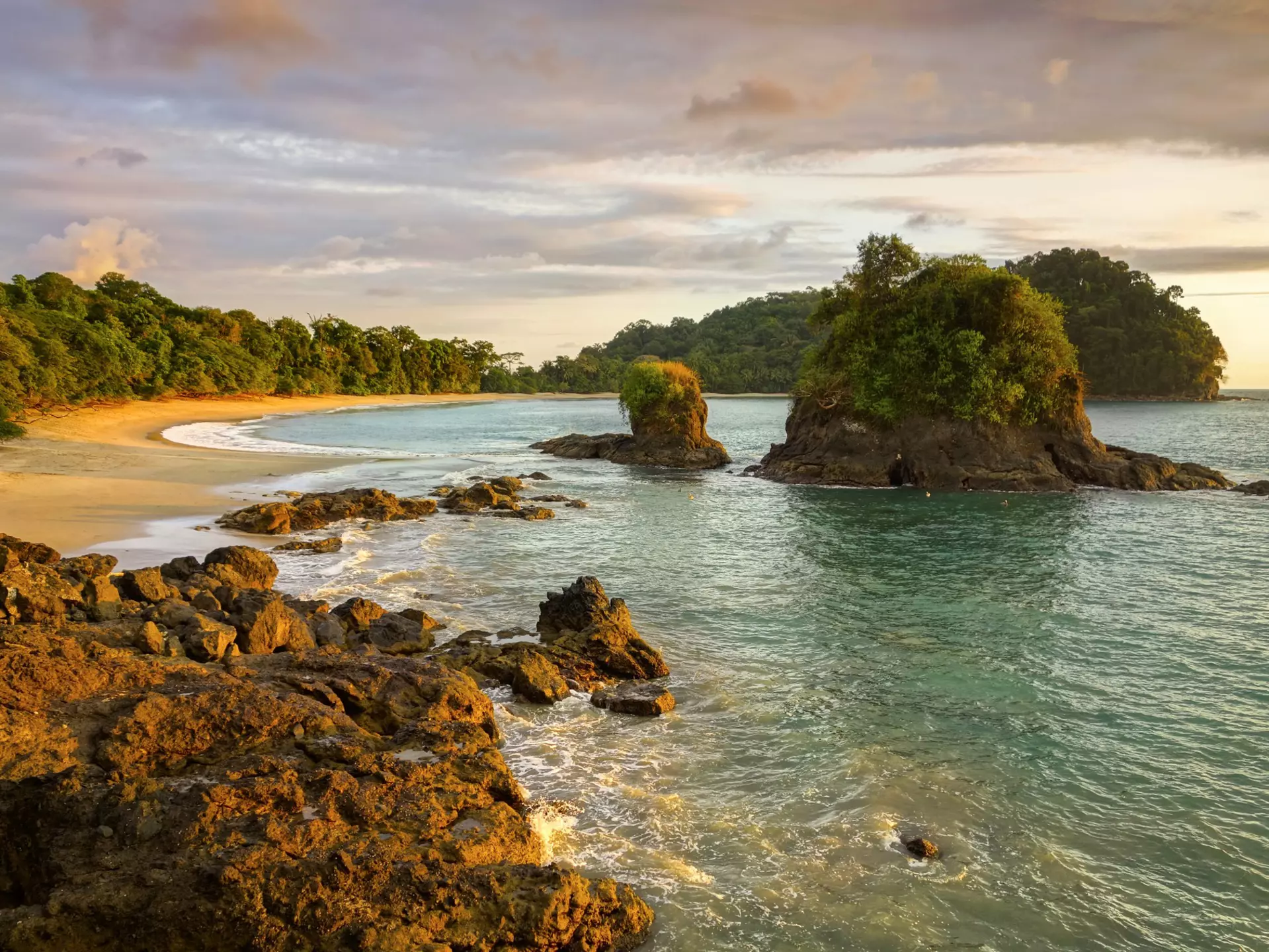 A secluded beach at the edge of jungle with nearby foliage-topped rocky islets.