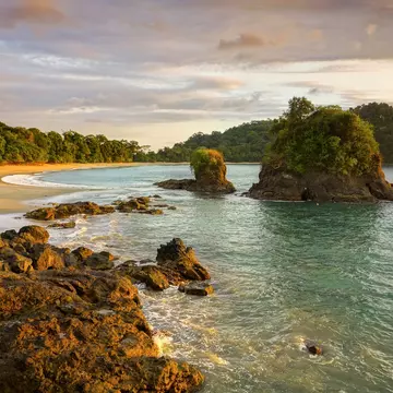A secluded beach at the edge of jungle with nearby foliage-topped rocky islets.