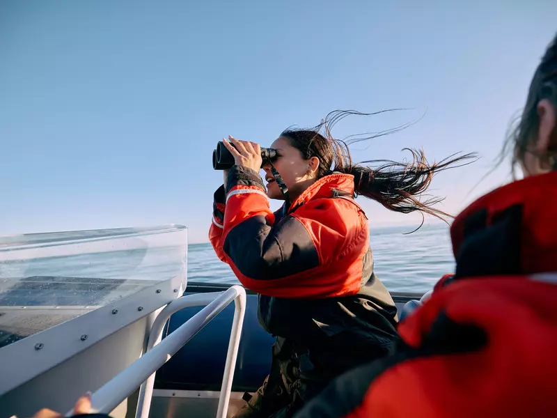 A woman in an orange jacket on a boat uses binoculars to look for whales off the coast of British Columbia
