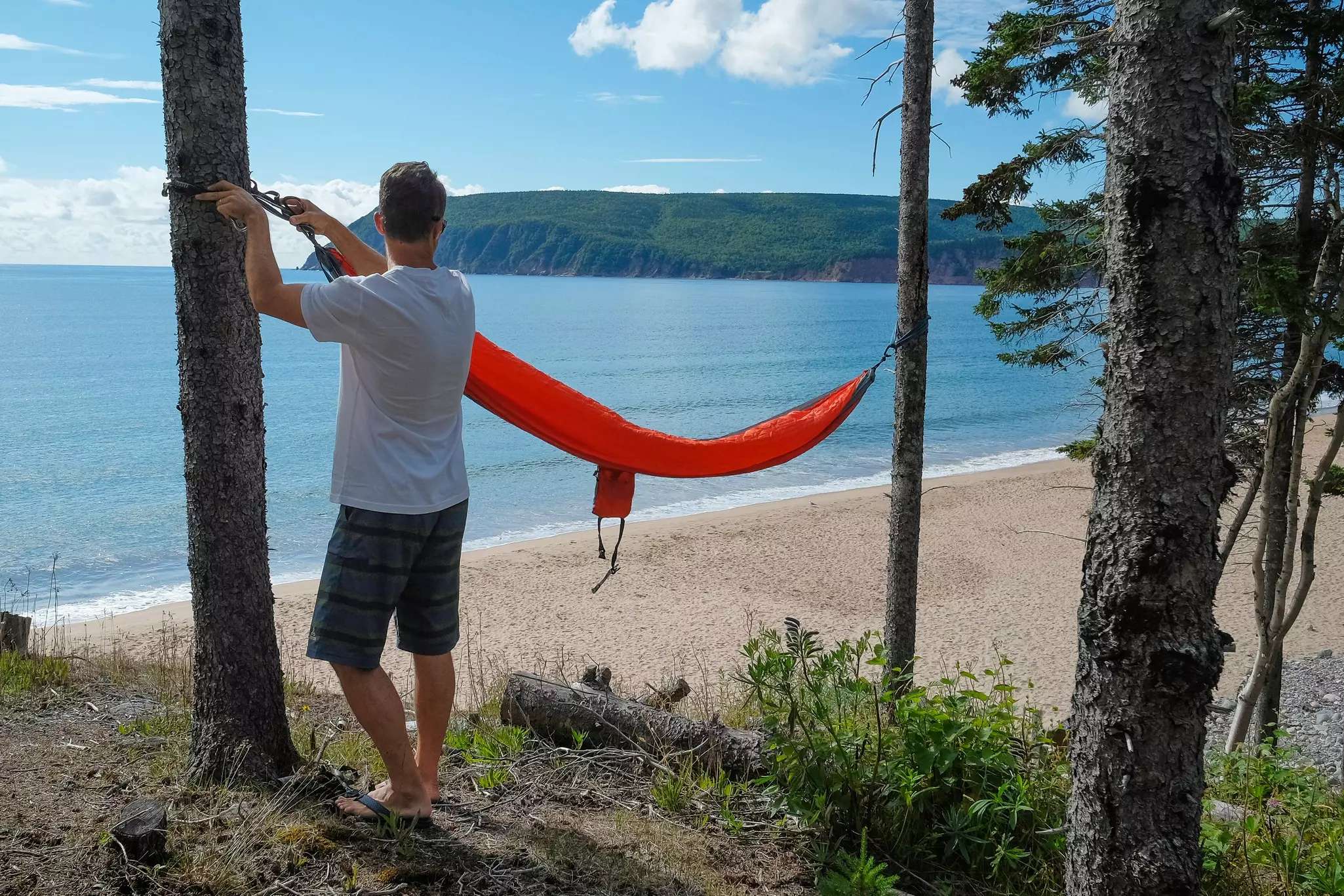 Man setting up a red hammock tied to two trees on the beach.