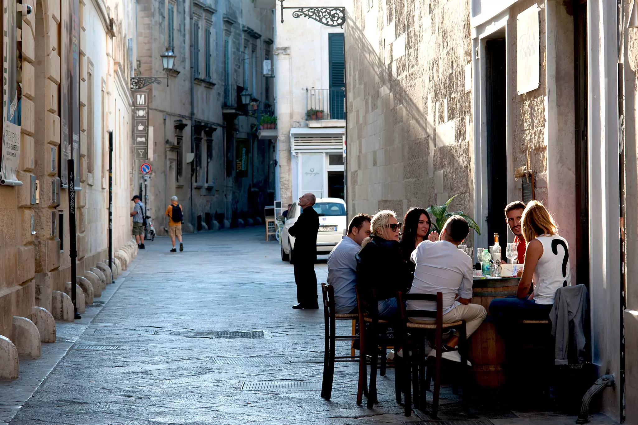On a narrow street in a historic town in Italy, people sit at a table with clear glasses in front of a cafe.