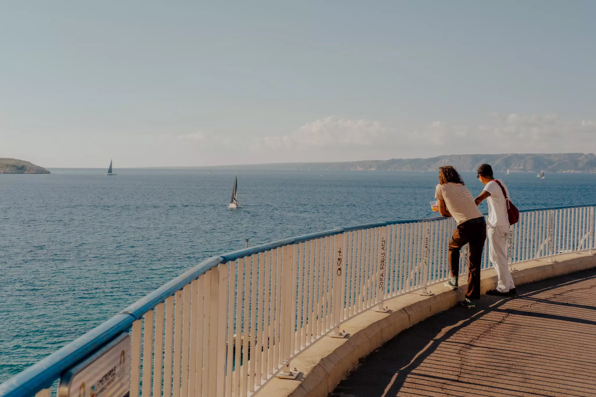 Two people stand against a railing looking out into the sea in front of them; there are sailboats in the water.