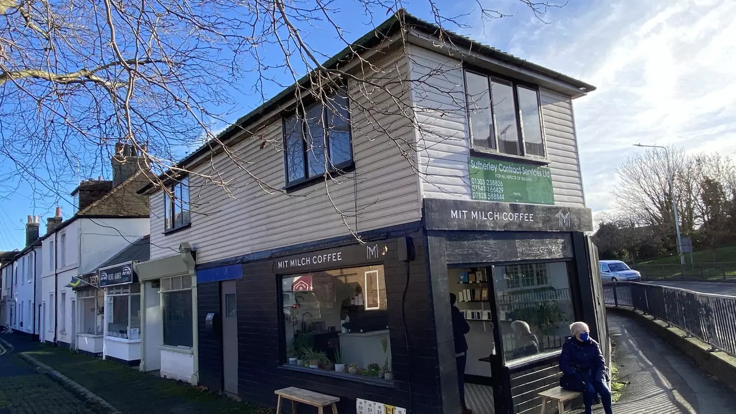 A person wearing a hat and warm coat, sitting on a bench outside Mit Milch coffee on a mostly clear day