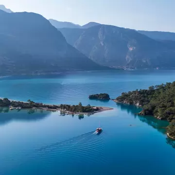 Boat leaving a wake across the blue lagoon at Ölüdeniz, a tree-lined sandbar and beach curving below forested mountains