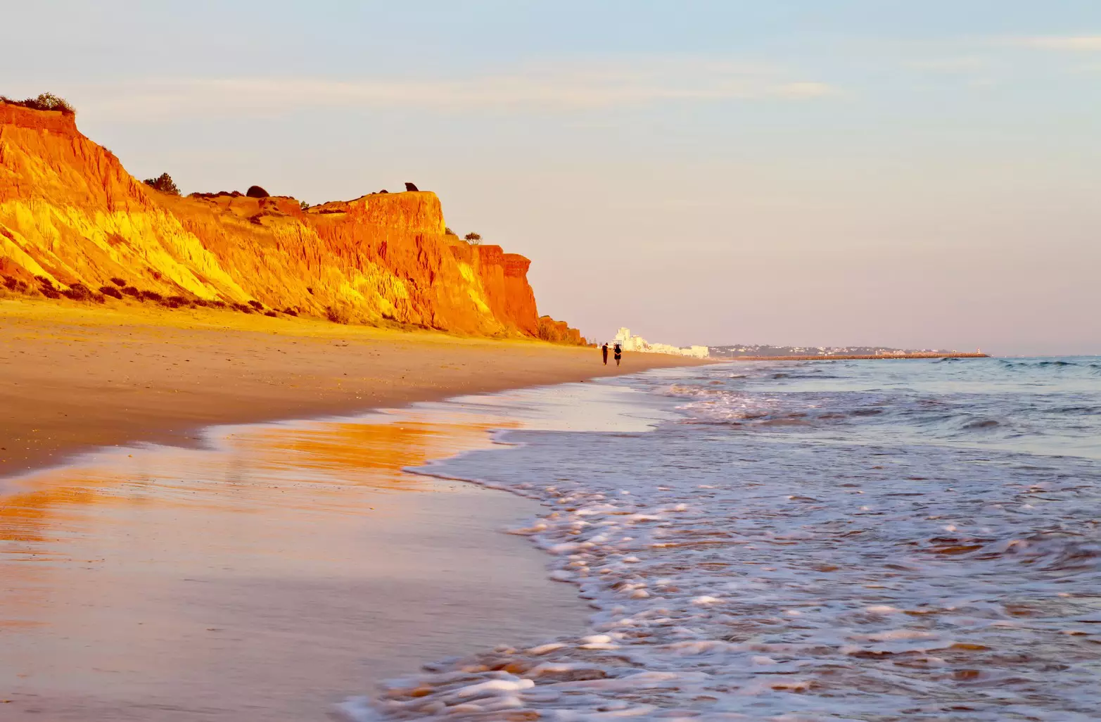 People walk at sunset along a beach backed with beautiful red cliffs.