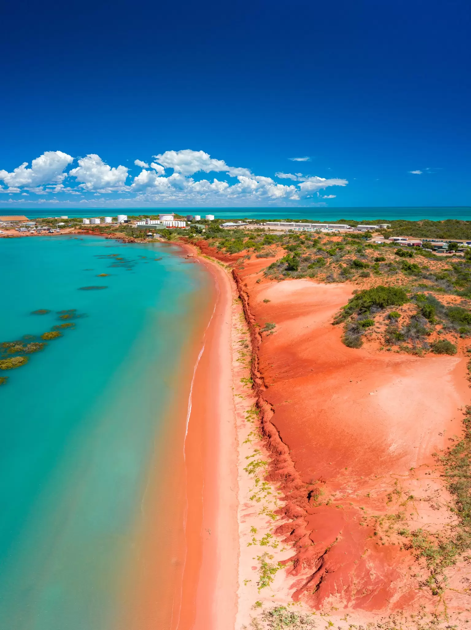 Turquoise ocean water is contrasted against a red beach with a town in the background. 