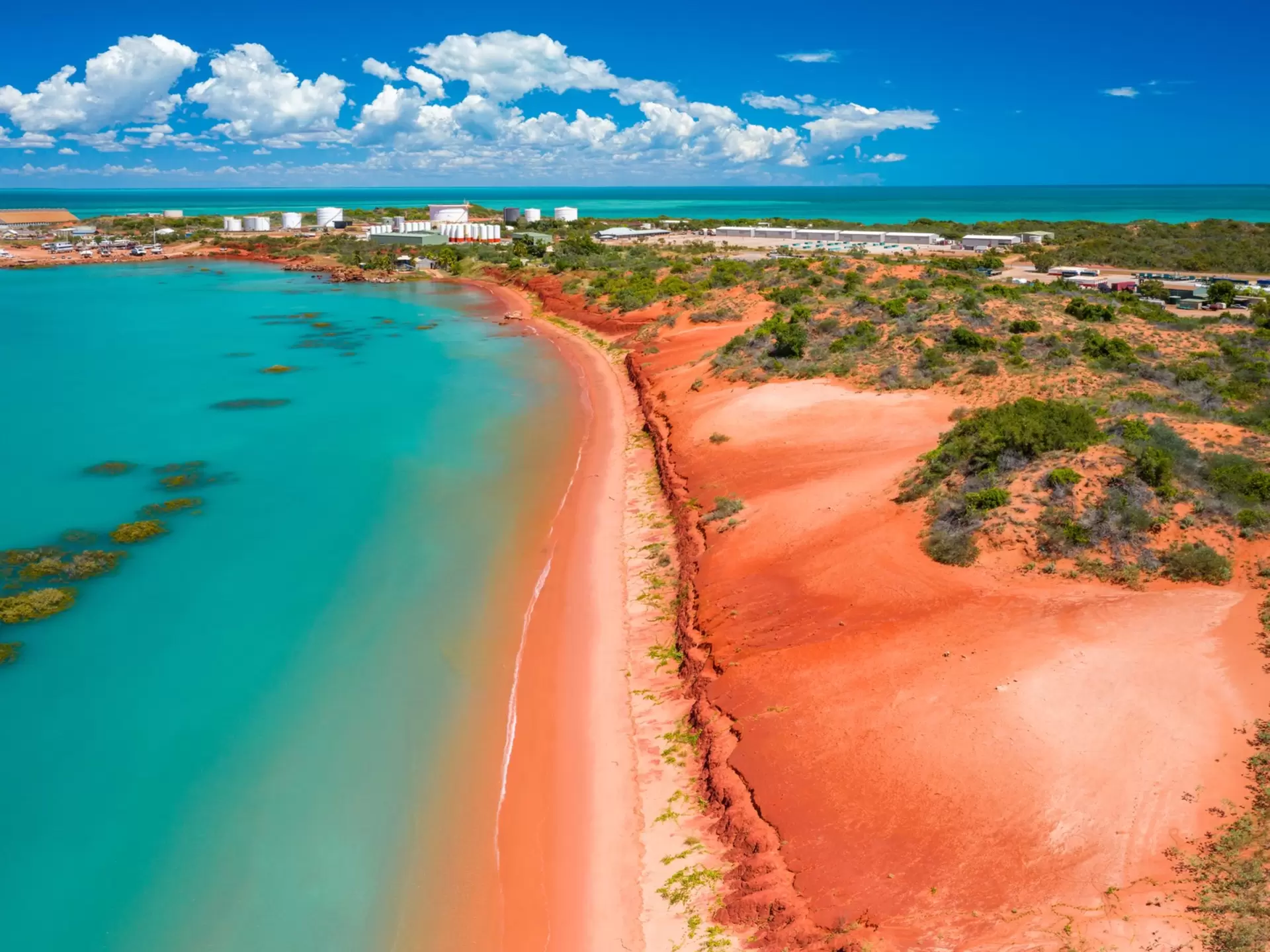 Turquoise ocean water is contrasted against a red beach with a town in the background. 