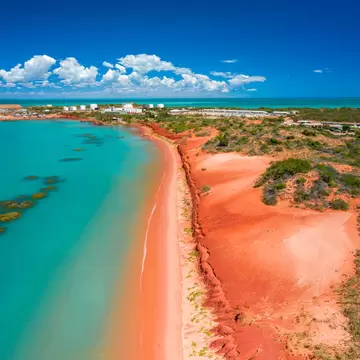 Turquoise ocean water is contrasted against a red beach with a town in the background. 