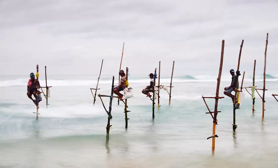 Four stilt fishermen sit on wooden poles above white surf off Weligama's coast in Sri Lanka under a grey overcast sky
