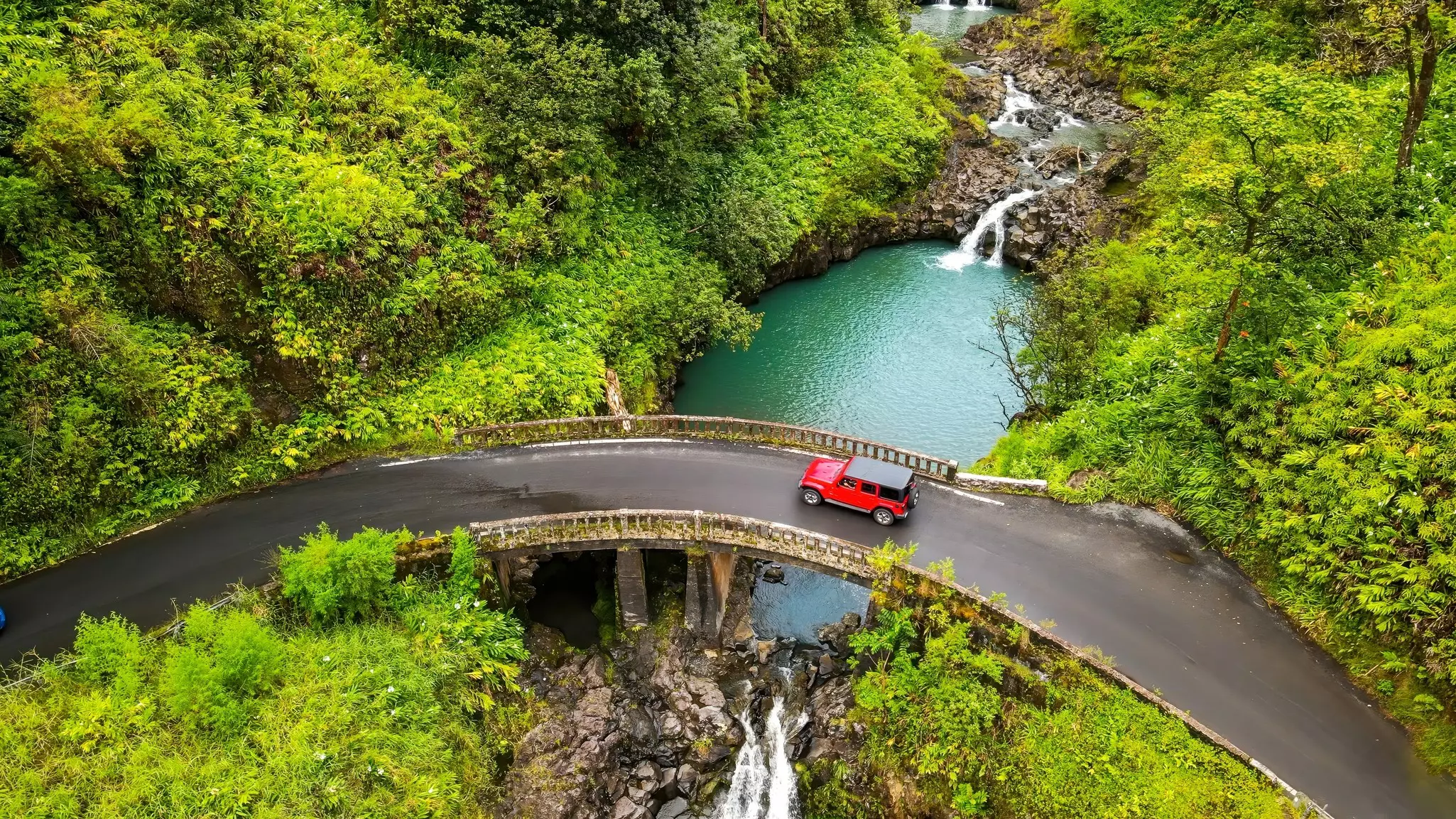 A red jeep travels a curved roadway on a stone bridge with a blue pool of water and small waterfalls below, surrounded by lush greenery on an overcast day.