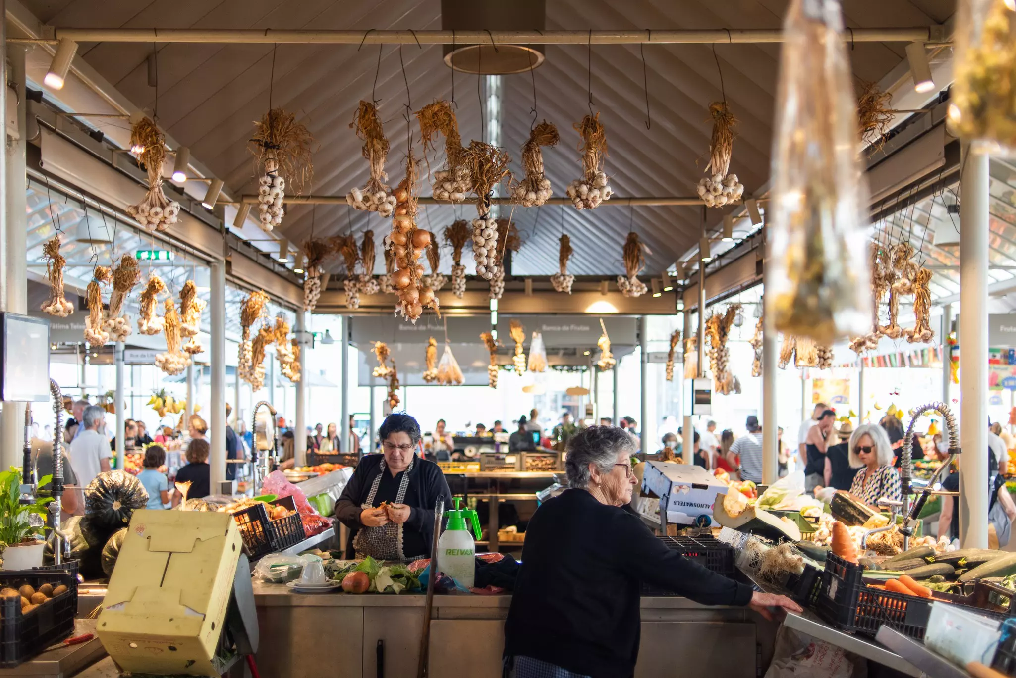 A produce stand at an indoor market has garlic and onions hanging from the frame of the structure.