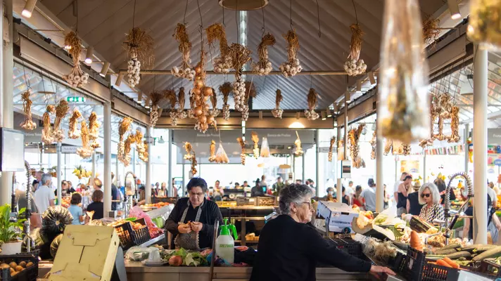 A produce stand at an indoor market has garlic and onions hanging from the frame of the structure.