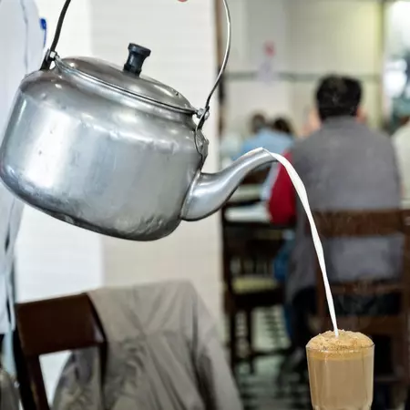 A waiter pours hot milk from a spouted silver kettle into coffee in a tall, clear glass.