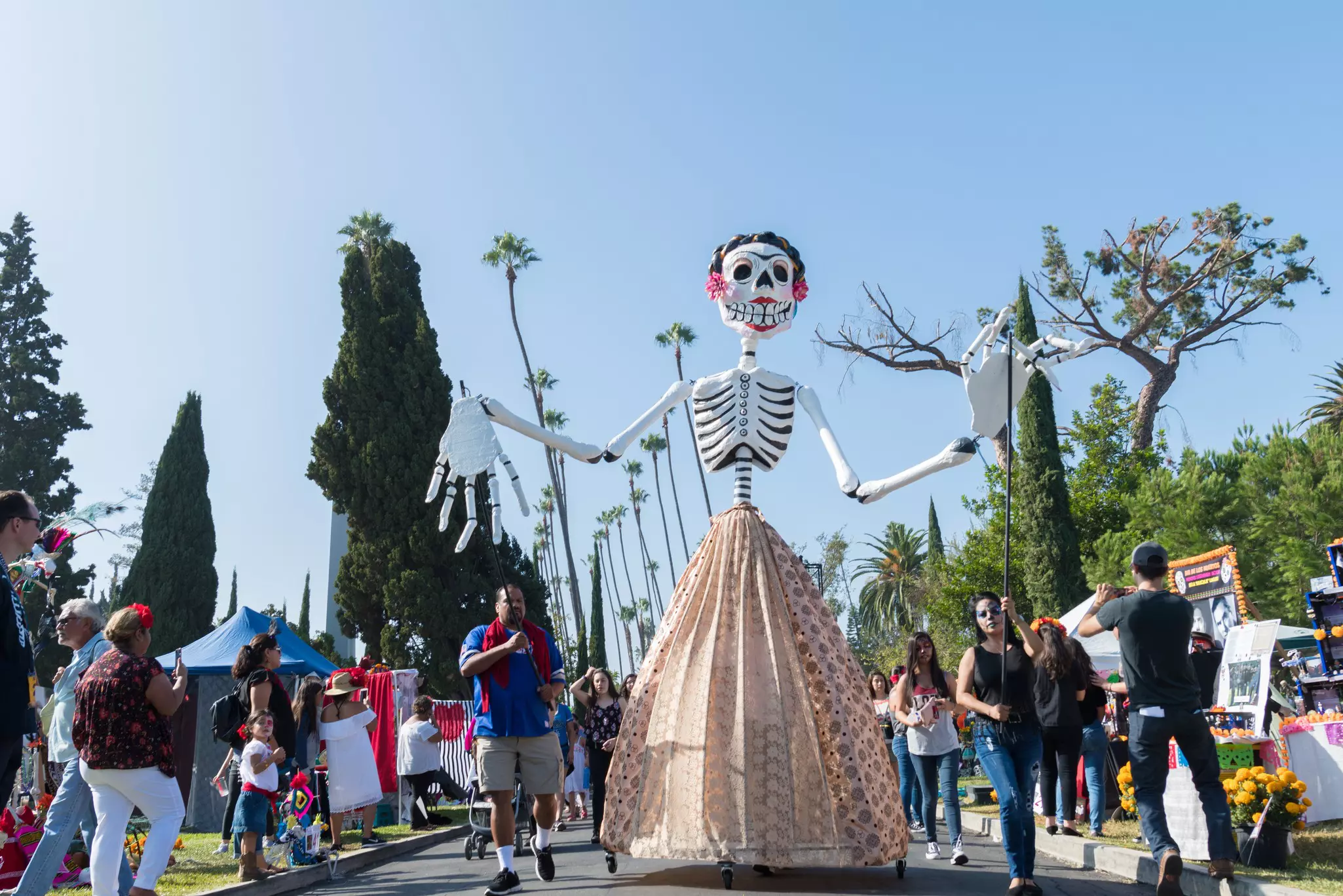 A large-scale skeleton puppet parades down a tree-lined pathway.