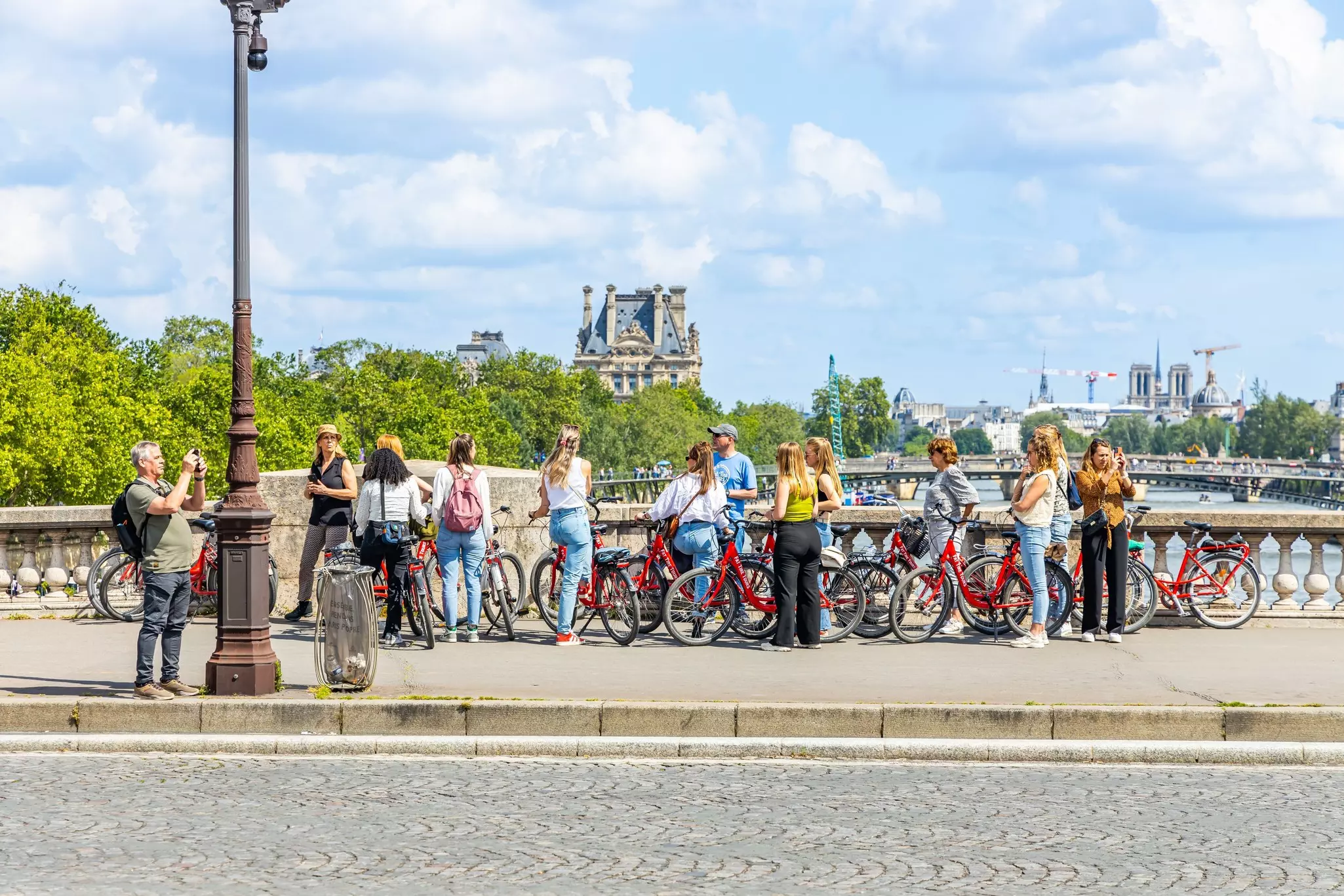 A group of people, each with a red bike, listen to a tour guide while pausing on a bridge over a river in a city.