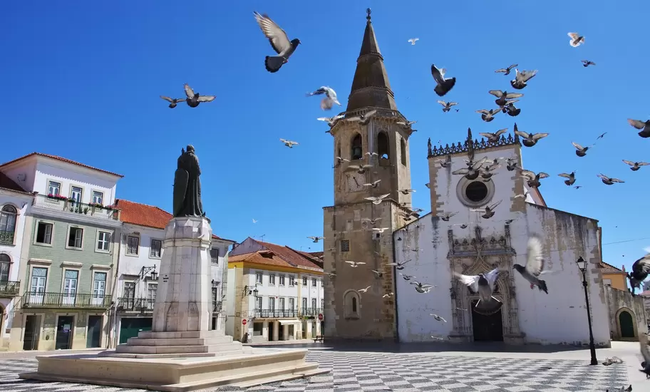 Pigeons fly over Praça da República in Tomar, Portugal, by the Igreja de São João Baptista bell tower and Gualdim Pais statue