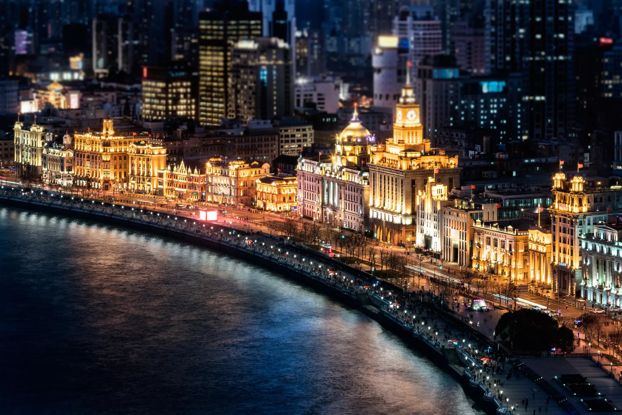 An aerial view of a riverbank by night, with elegant buildings illuminated to dramatic effect, the light reflecting off the water.