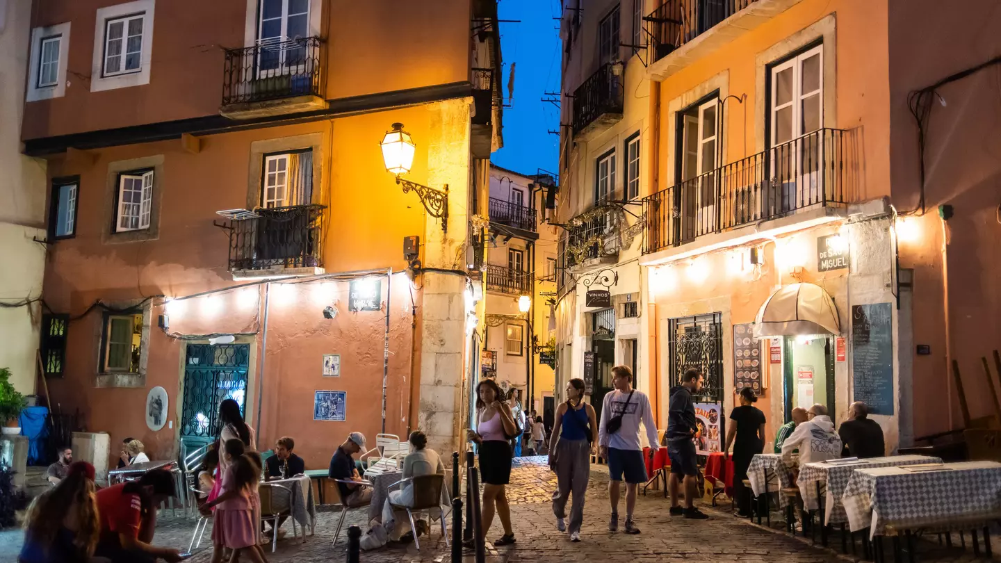 People walk down a narrow cobbled street in a city in the evening, past tables at outdoor restaurants.