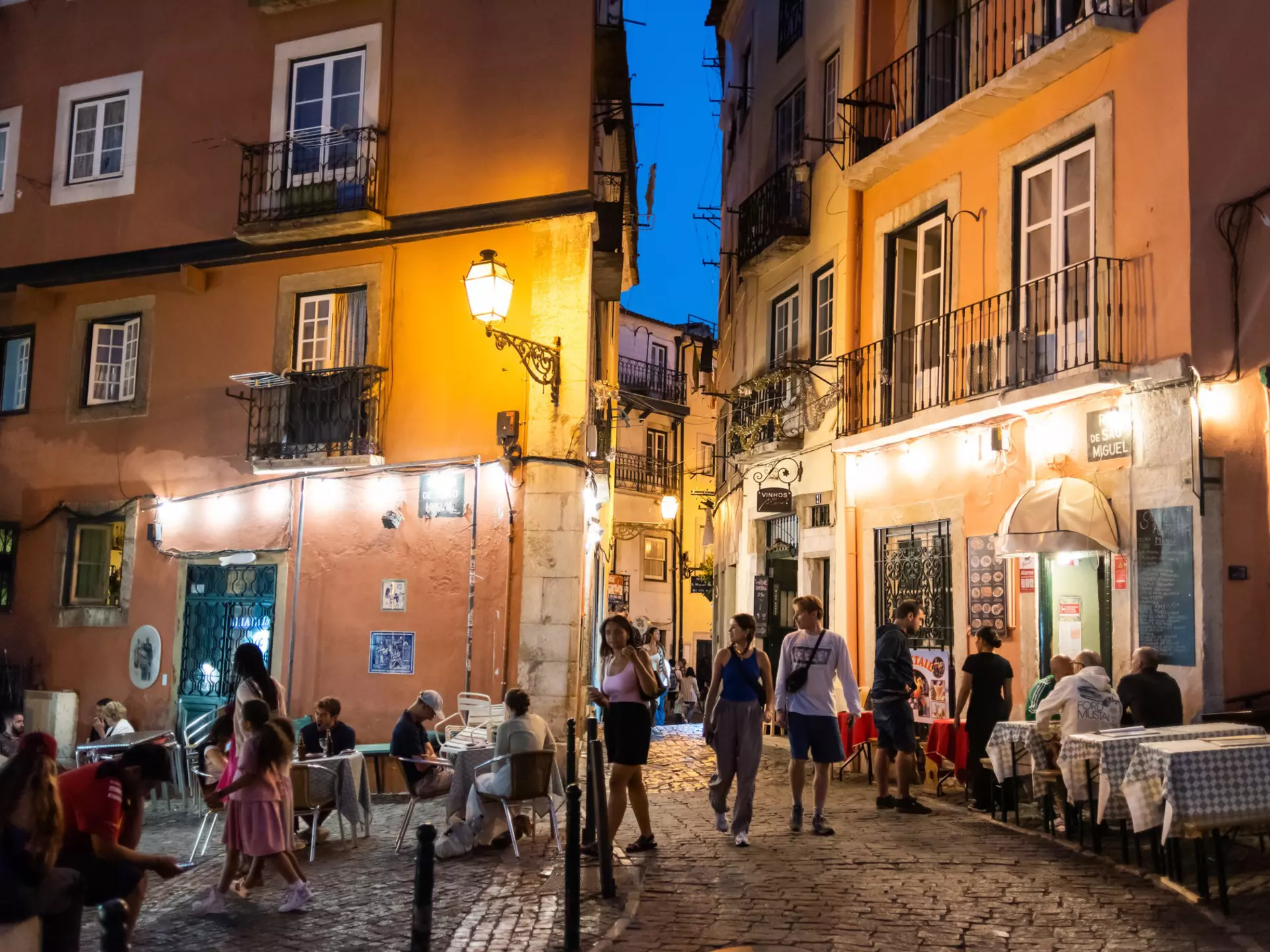 People walk down a narrow cobbled street in a city in the evening, past tables at outdoor restaurants.