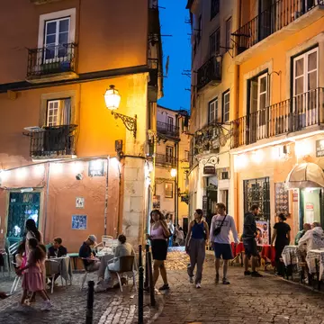 People walk down a narrow cobbled street in a city in the evening, past tables at outdoor restaurants.