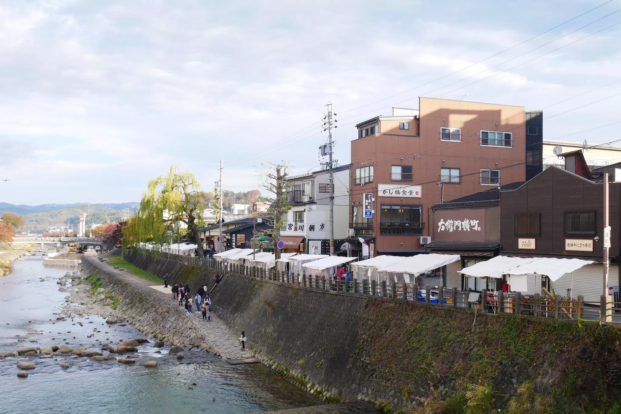 A market of white tent stalls alongside brick buildings and a riverfront embankment  on a partly sunny day.