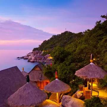 Lit thatched-roof huts on large boulders beside a jungle hillside, overlooking a calm sea under a pink evening sky.