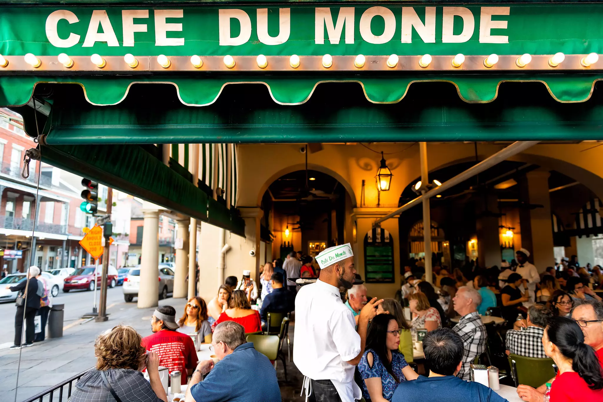 Dozens of people sit at tables at an outdoor seating area covered with a green awning. A server in a white uniform and cap takes orders.