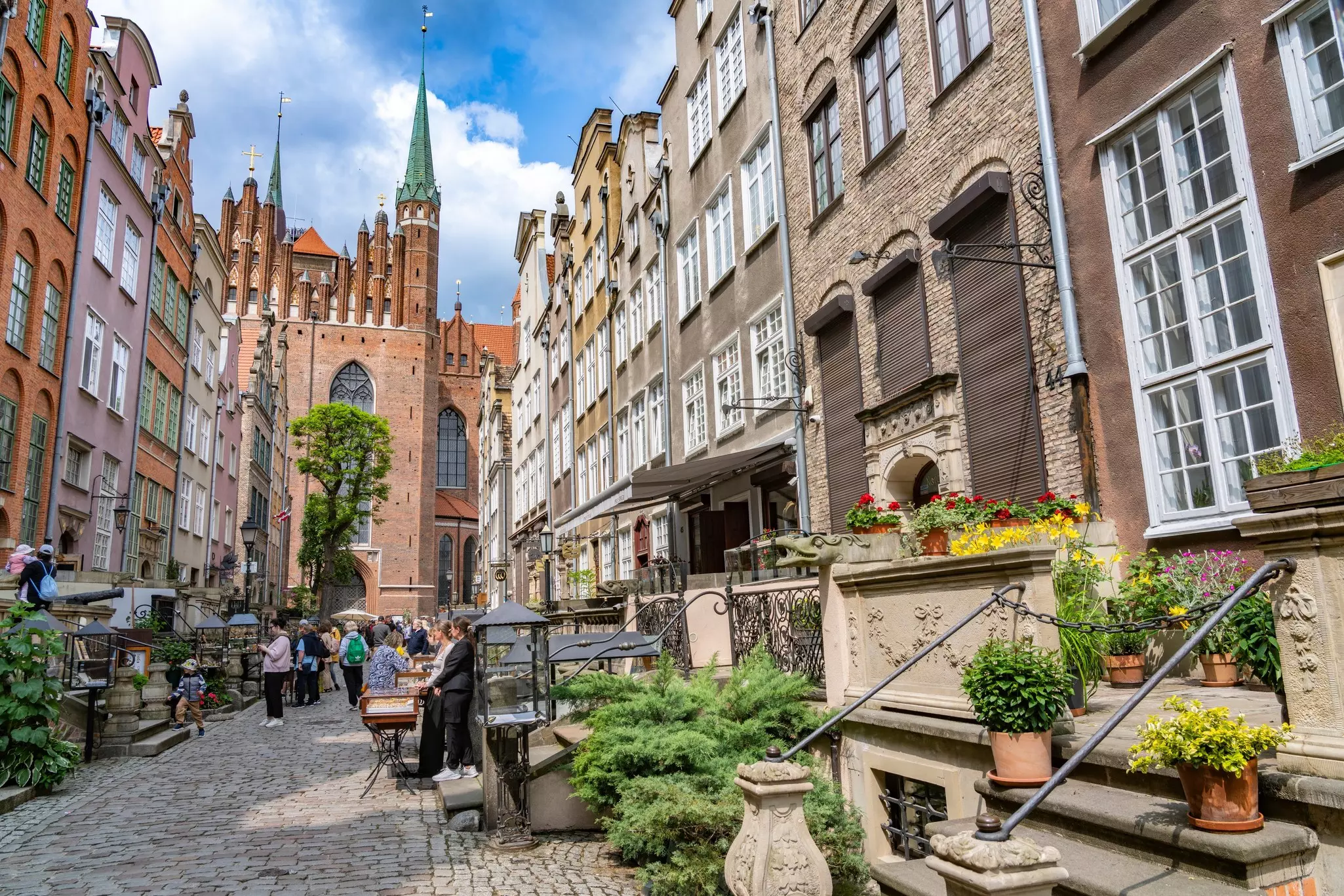 People on a stone pedestrian street near old stone buildings with filled flowerpots in front of them and on their steps