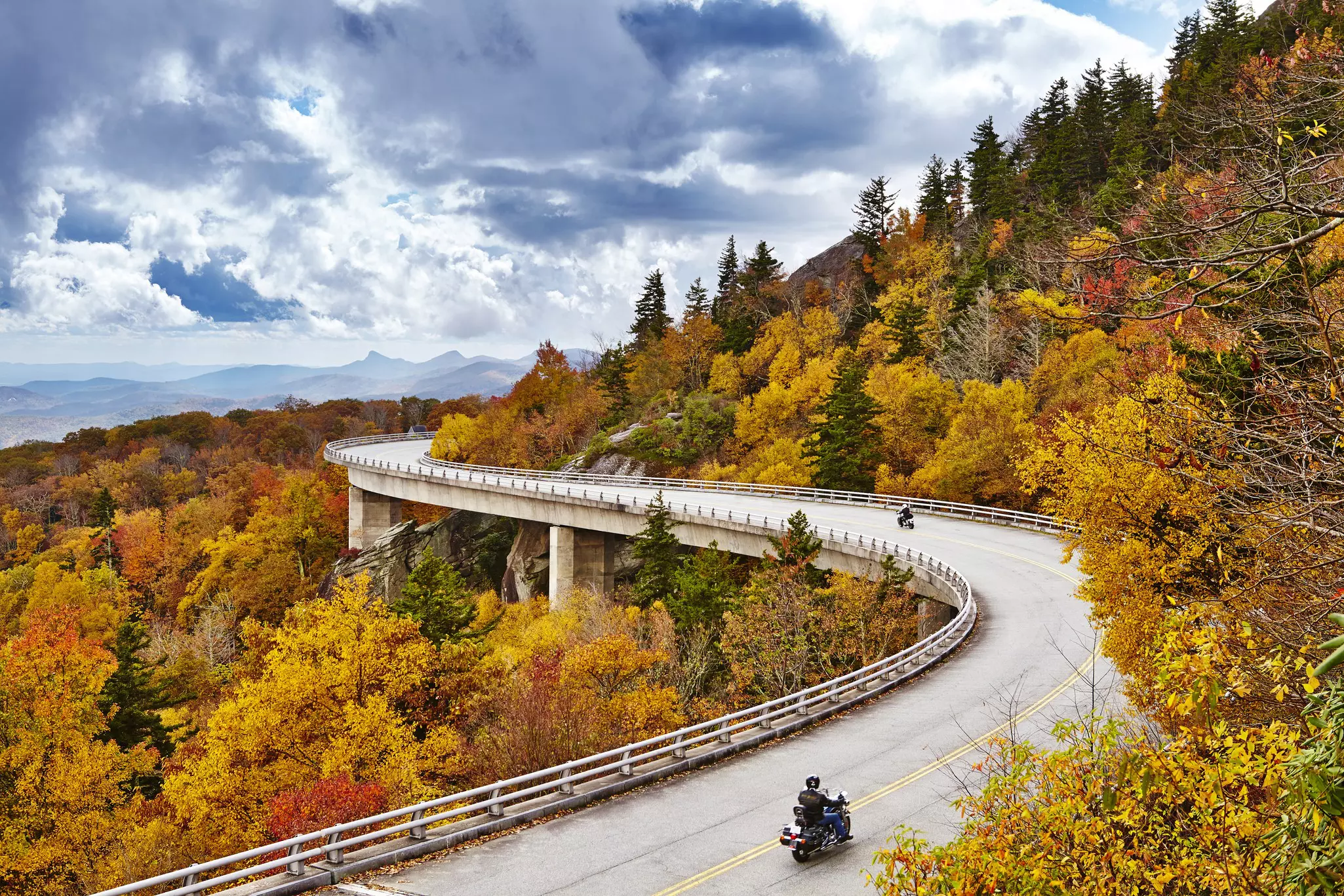 Two motorcycles on a weaving road raised above dense woodland that glows gold, orange, yellow and red during fall.