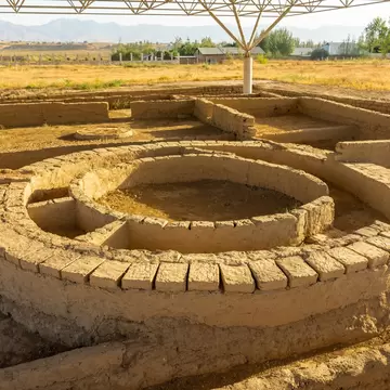 Low mudbrick walls of an excavated settlement, including a large circular structure, under a modern canopy with hills behind.
