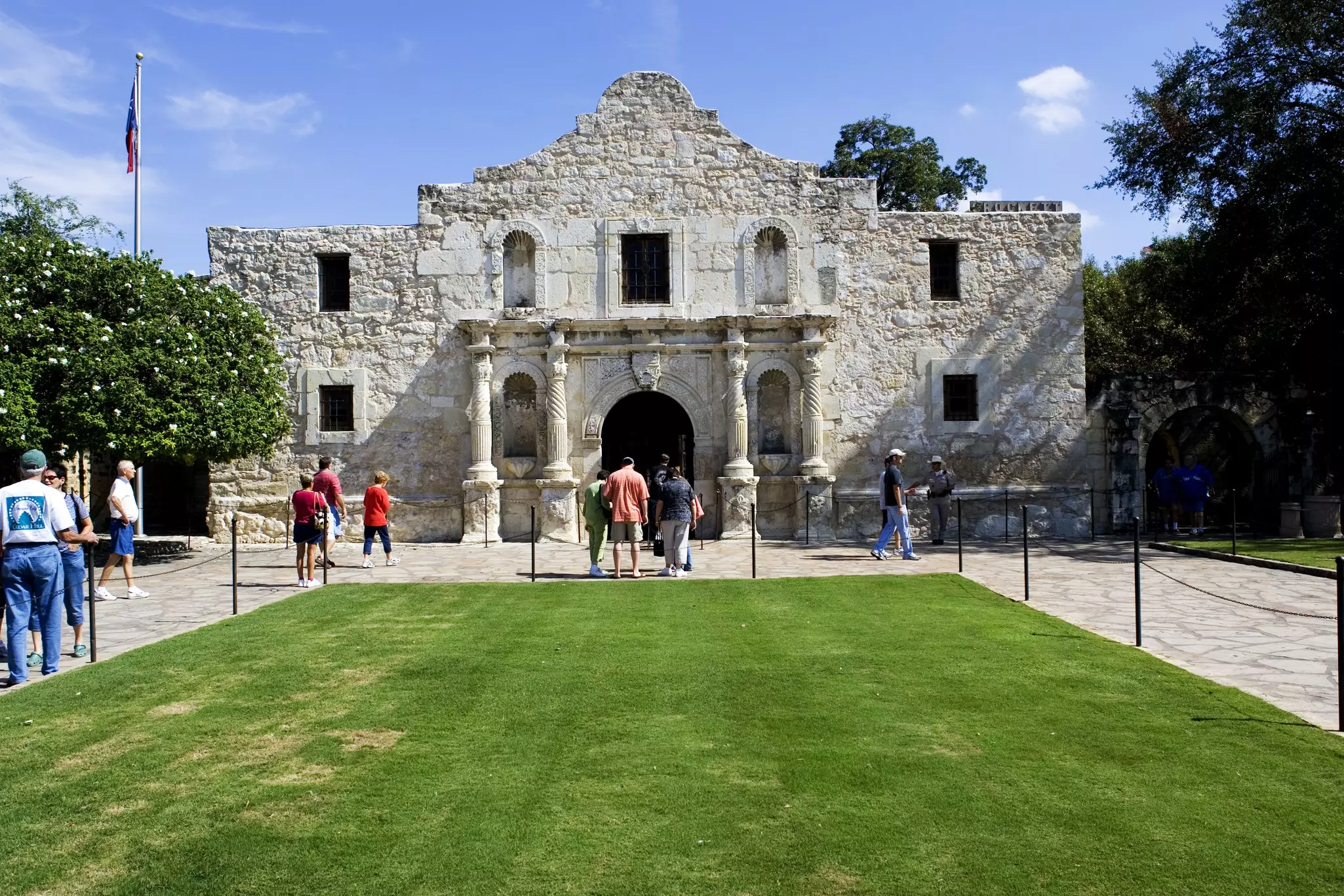 A neat lawn leads to a low-rise historic stone building with tourists waiting to enter.
