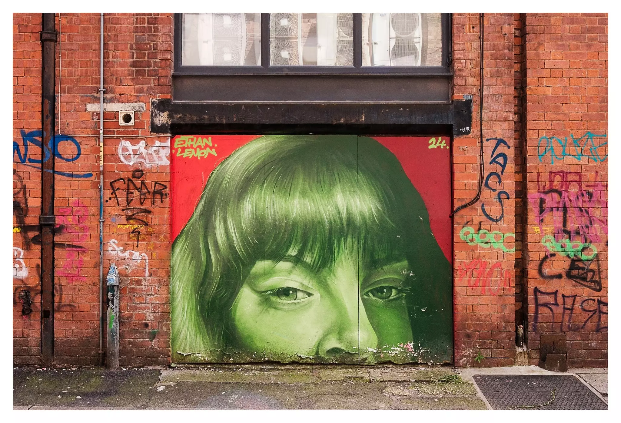 Close-up of brick wall with graffitti lettering and a mural picturing the top half of a woman's face in green paint.