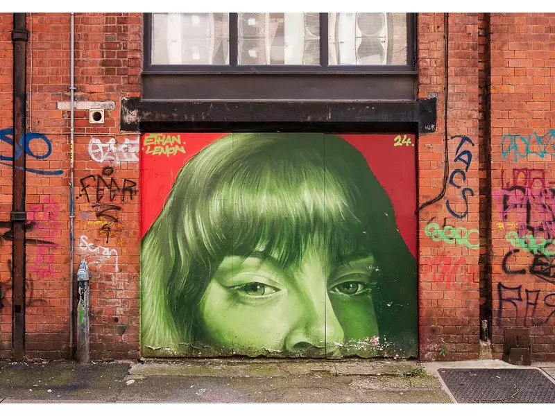 Close-up of brick wall with graffitti lettering and a mural picturing the top half of a woman's face in green paint.