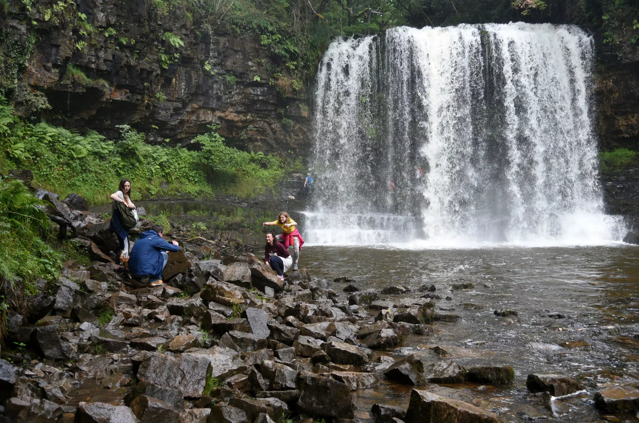 A group of hikers poses on rocks in front of a wide waterfall.