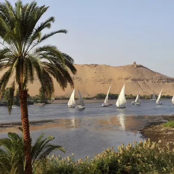 Sailing boats with white sails cruise along a wide river. The tall riverbank opposite has tombs cut into the rock face.