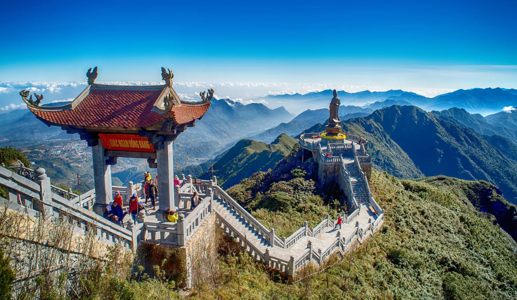 A red-roofed archway stands over a staircase leading down to the Great Buddha statue at Mt Fanispan in Vietnam, with blue haze over the mountain range behind it
