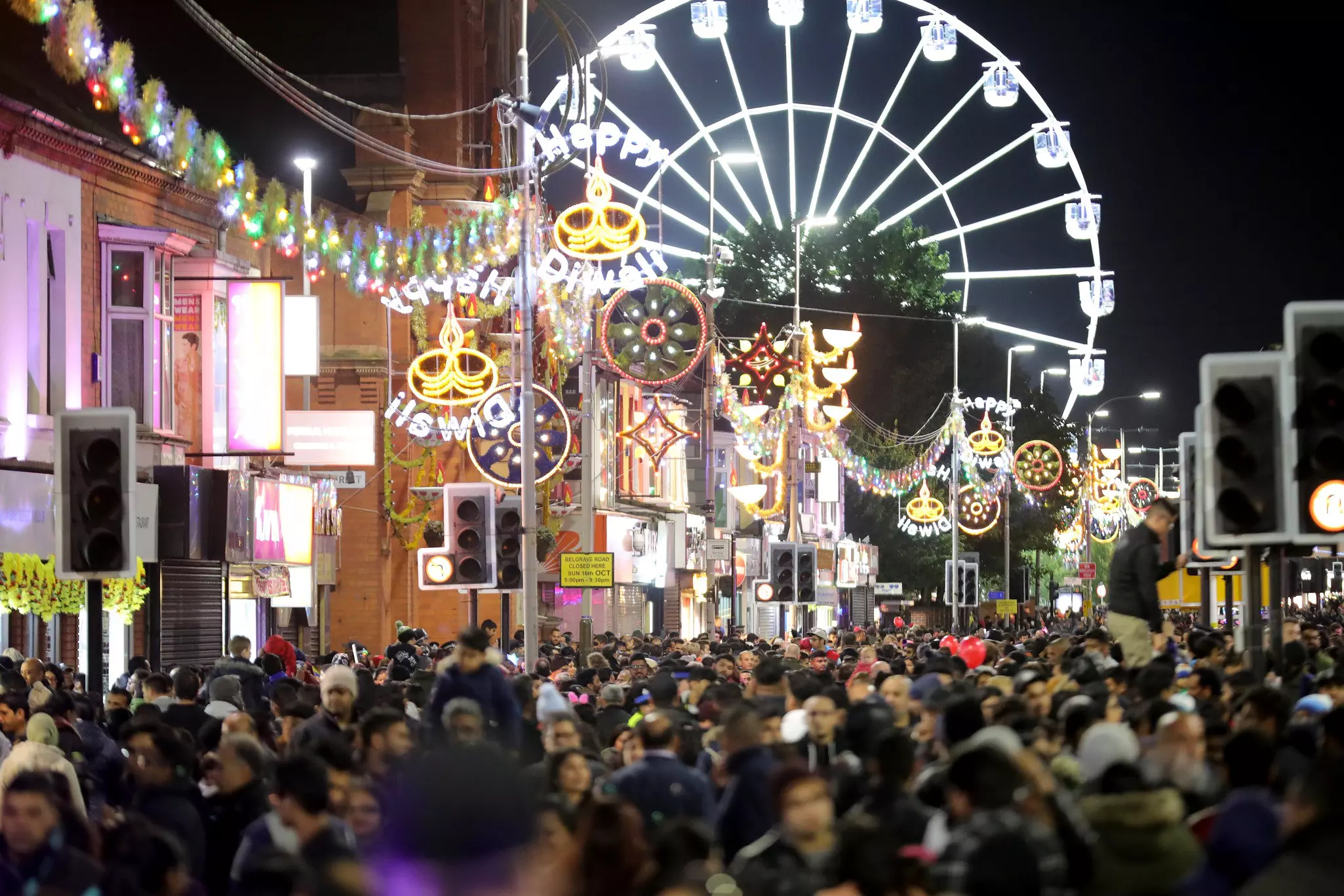 A dense crowd of people throngs a street in a city after dark, the scene illuminated by bright lights. A large Ferris wheel is seen in the background.