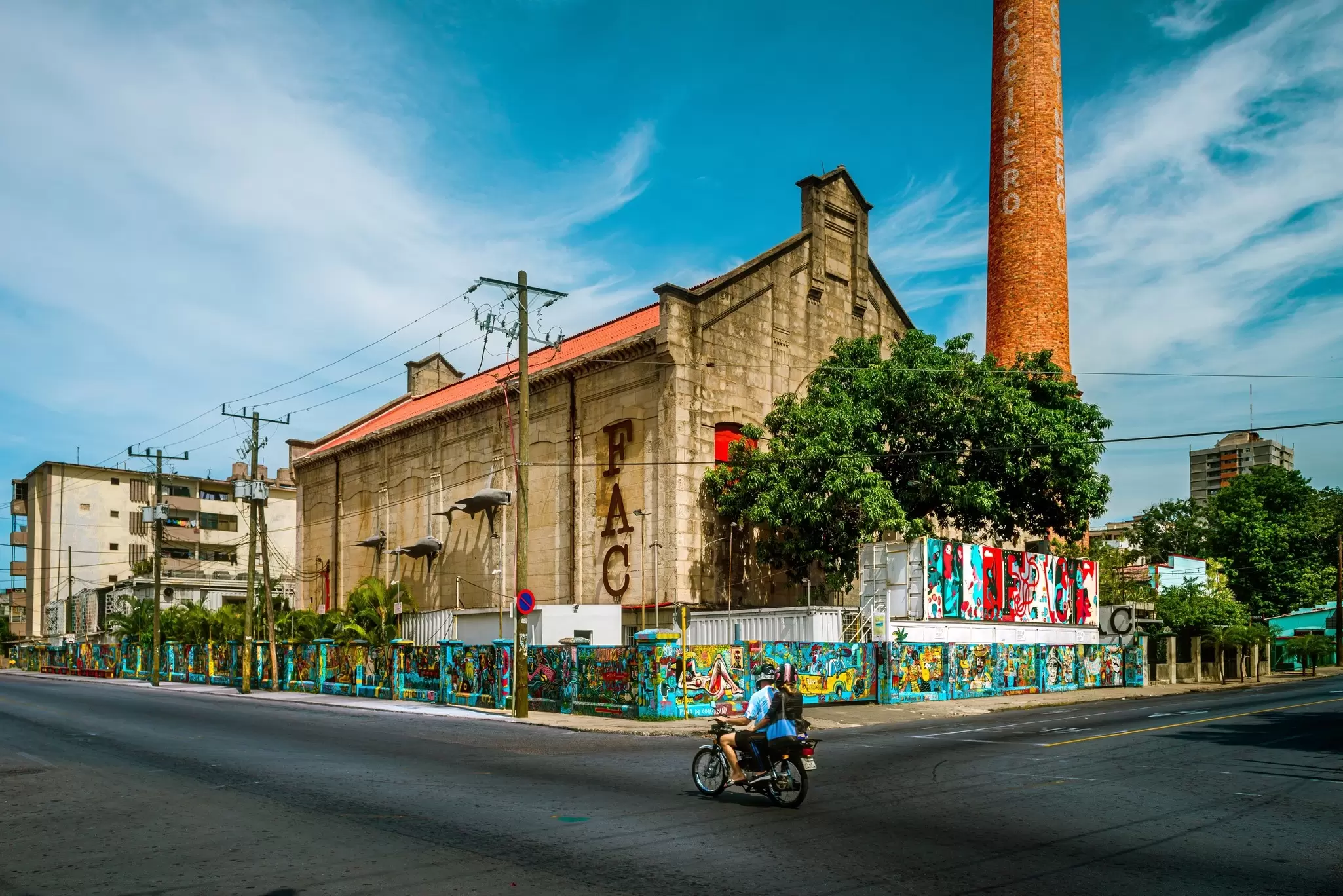 A young couple ride and small motorbike passing by a famous old warehouse turn into an artistic place F.A.C. Fabrica de Arte Cubano in El Vedado, Cuba.