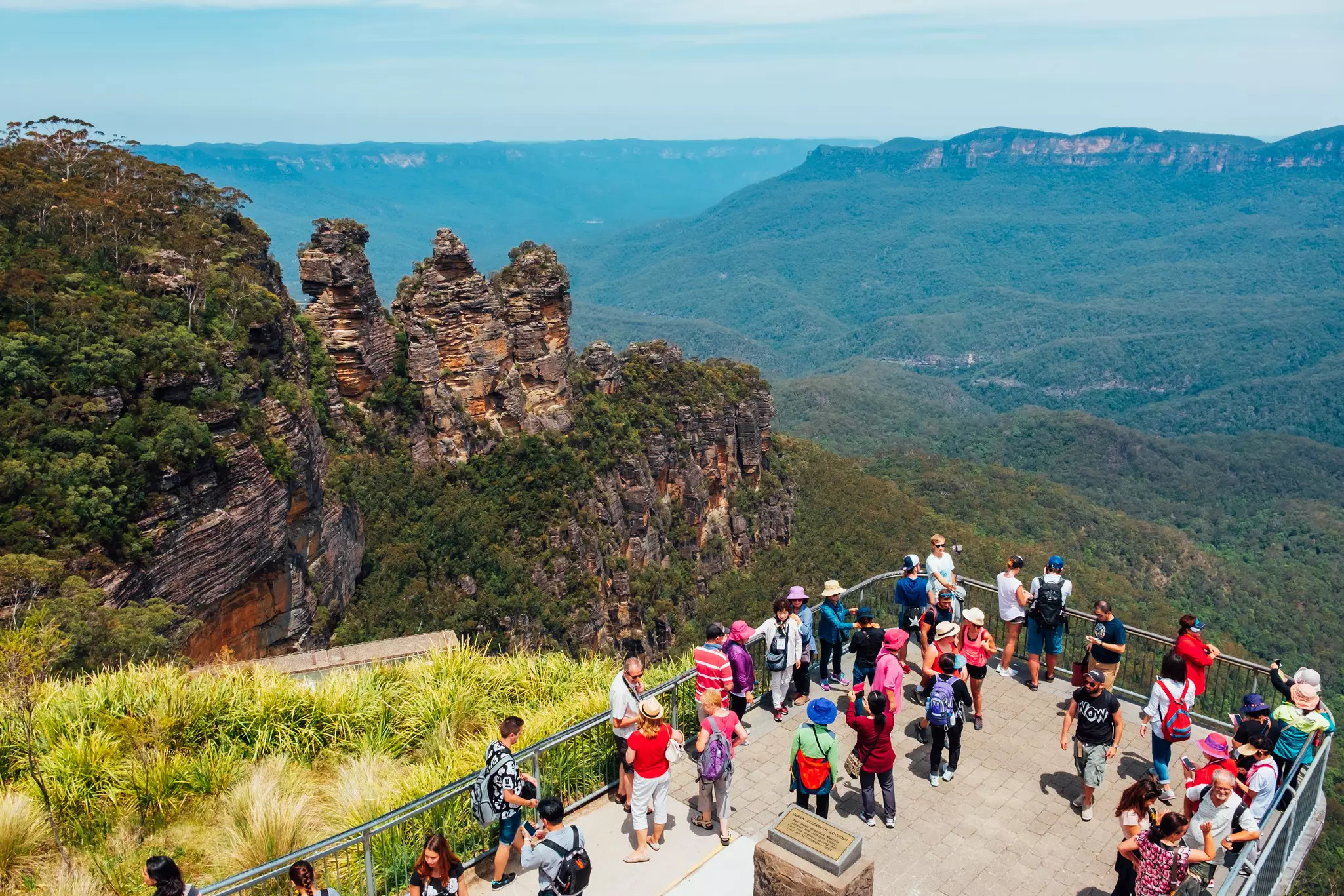 A group of tourists at a lookout point take photos of the scenery. Rolling hills covered in thick forest stretch into the distance. A rock formation of three distinct pillars stands nearby.