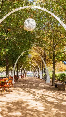 White arches stretch over a path with orange tables and chairs to the left and shady trees lining the path