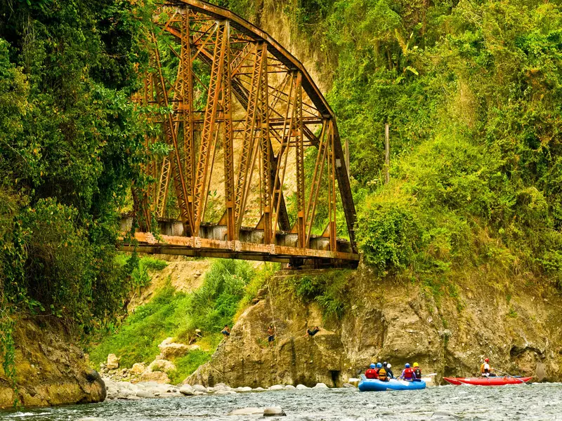 Rafters paddling under an old railroad bridge with kids swinging from a rock