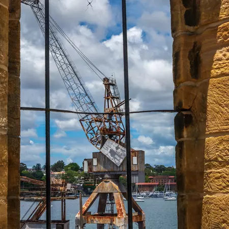 A disused crane along a waterfront viewed from a window inside an old building on a sunny day with puffy clouds in the sky..