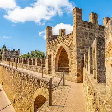 A crenellated sandstone gate tower with a pointed arch, seen along the parapet walk of a medieval town wall above rooftops.