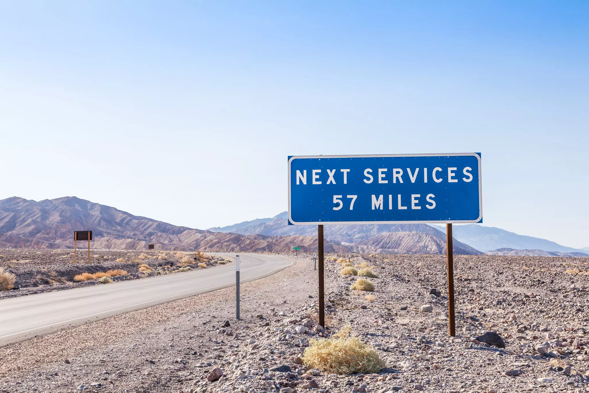 A blue and white sign beside a desert road that states "next services 57 miles"
