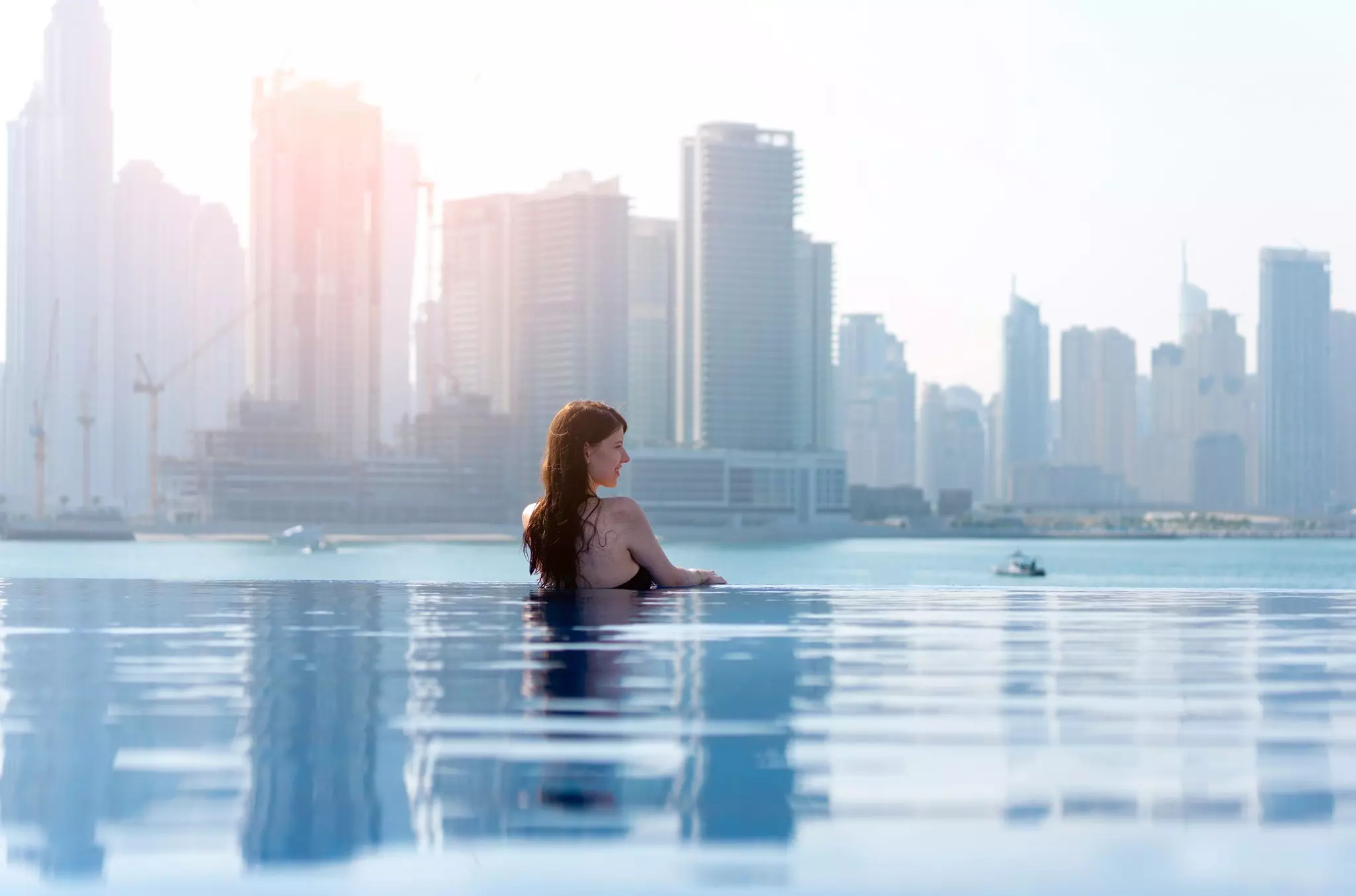 A woman is pictured in an infinity pool, with the skyline of a city behind her, across a bay.
